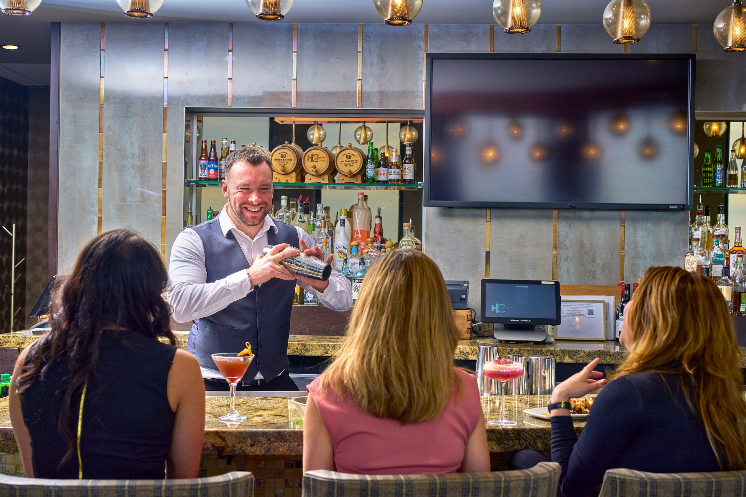 Bartender smiling while shaking a cocktail with 3 women seated at The Houston Club bar